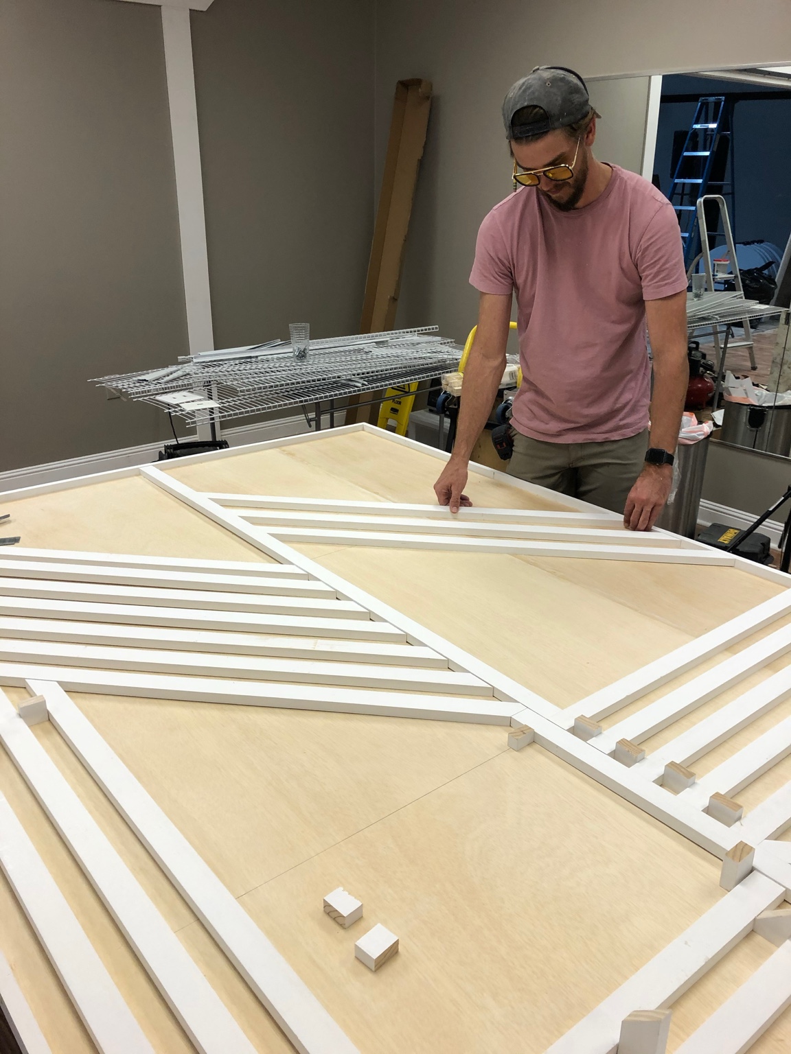 Carefully positioning white trim in a geometric pattern on a large wood panel during construction.